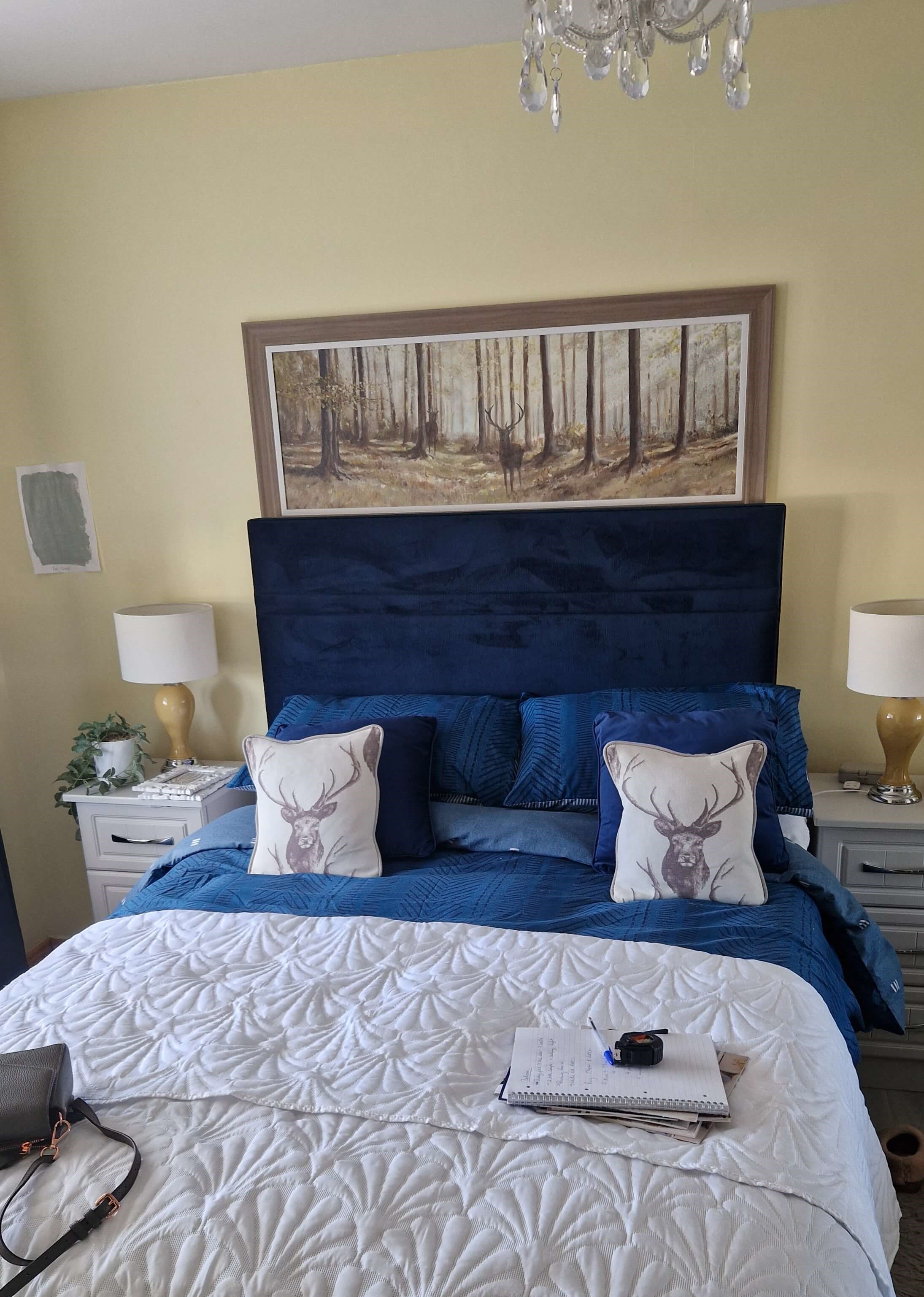 a bedroom with lemon yellow walls, a bed with navy, velvet headboard. Two white, wooden side tables with lamps on each one with a golden colour stand and white shade. a Picture of a deer in the forest is above the bed.